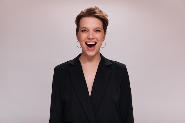 Free photo portrait of brown eyed lady in dark jacket on isolated background. attractive short-haired woman in black suit laughs and looks into camera on white backdrop