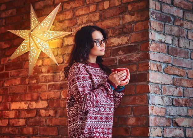 Portrait of brunette female dressed in a red sweater drinks coffee over the wall of a brick.