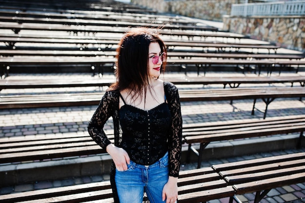 Free photo portrait of brunette girl in pink glasses wear on black posed outdoor on sunny day against row of benches