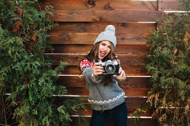 Free photo portrait brunette girl with red lips and long hair in winter clothes having fun with camera on wooden .