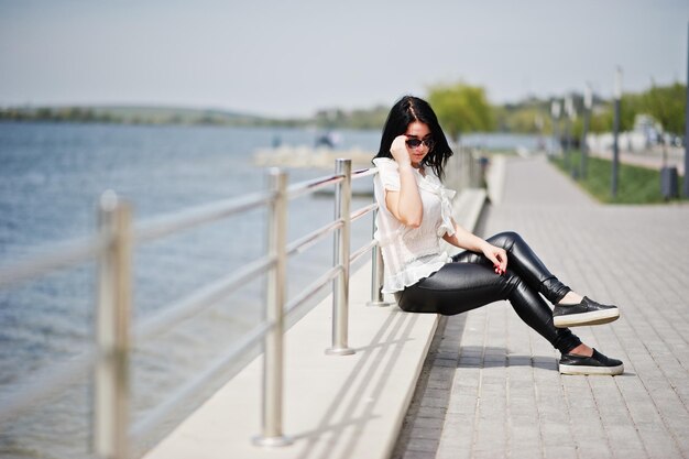 Portrait of brunette girl on women's leather pants and white blouse sunglasses against iron railings at beach