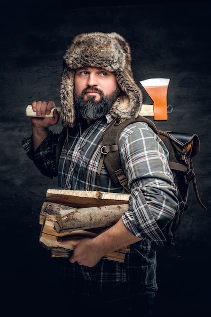 Portrait of brutal bearded hunter male dressed in a fleece shirt and a hat from a fur holds an axe on his shoulder.