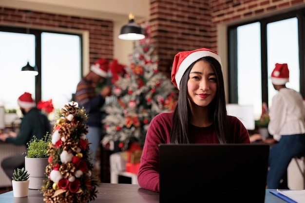 Free photo portrait of business woman working in christmas season in startup office decorated with festive xmas ornaments and lights. asian company worker using laptop during seasonal celebration.