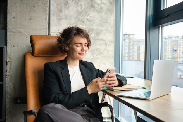 Free Photo portrait of businesswoman resting in her office in front of window using mobile phone sitting with