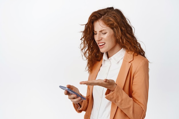 Free photo portrait of businesswoman staring confused at mobile phone arguing about something on smartphone standing over white background