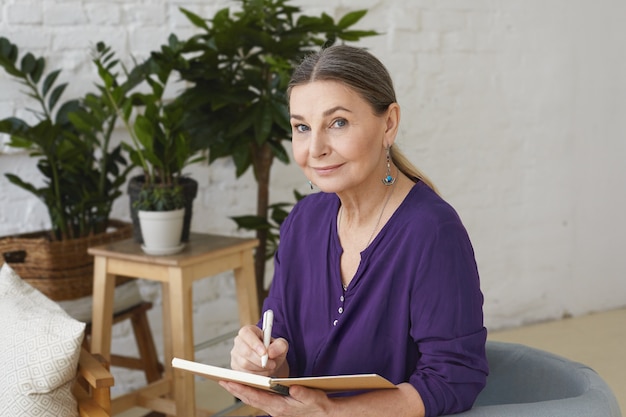 Portrait of busy modern 50 yeard old middle aged woman in violet shirt writing in copybook, making plans, looking with positive friendly smile, sitting on chair, surrounded with green plants