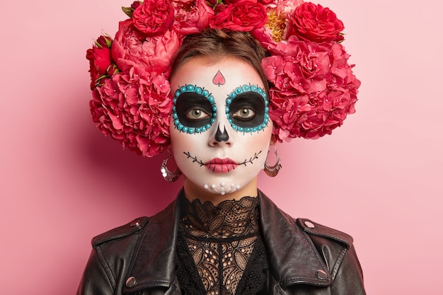 Free photo portrait of calm female celebrates day of death, has sugar skull makeup, dark circles near eyes, painted smile, thinks death is natural part of human cycle, wears traditional mexican attire.