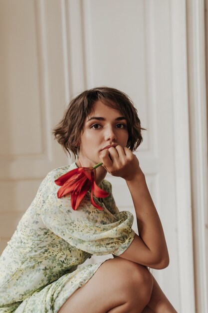 Portrait of calm young brunette short-haired woman in floral dress leans on knees, looks at front and holds red flower