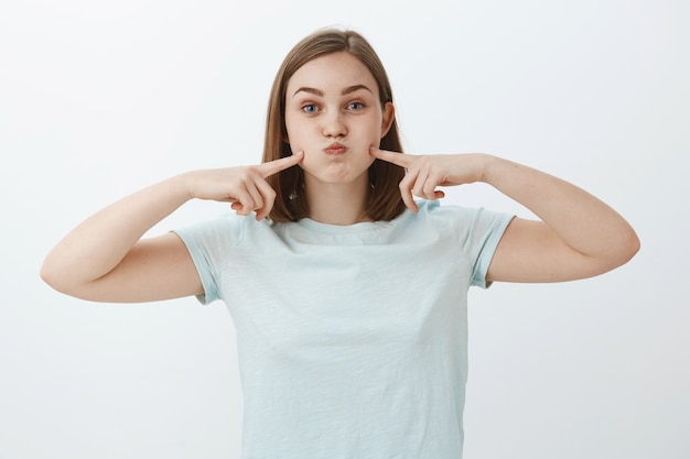 Portrait of carefree cute european female in casual light-blue t-shirt pouting, holding breath and poking cheeks with index fingers having fun spending time over grey wall trying amuse herself