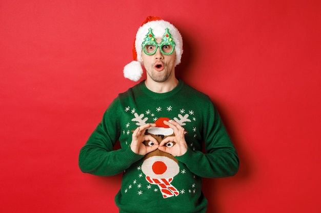 Portrait of carefree handsome man in santa hat and party glasses, making fun of his christmas sweater, looking happy over red background