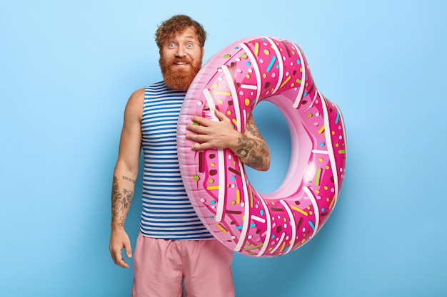 Portrait of carefree smiling red haired man posing with donut pool floaty