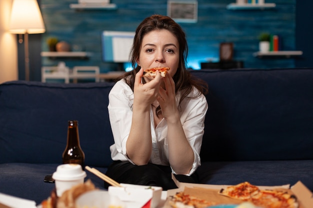 Portrait of caucasian female sitting on sofa eating slice of pizza enjoying takeaway food home deliv...