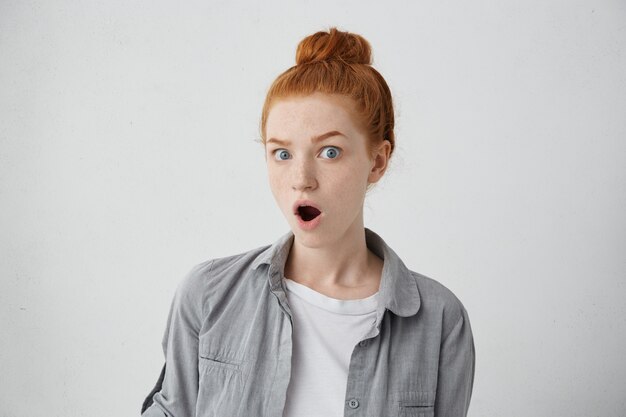 Portrait of Caucasian girl with blue eyes, freckles and ginger hair in knot having terrific look with opened mouth and eyes wearing casual clothes.