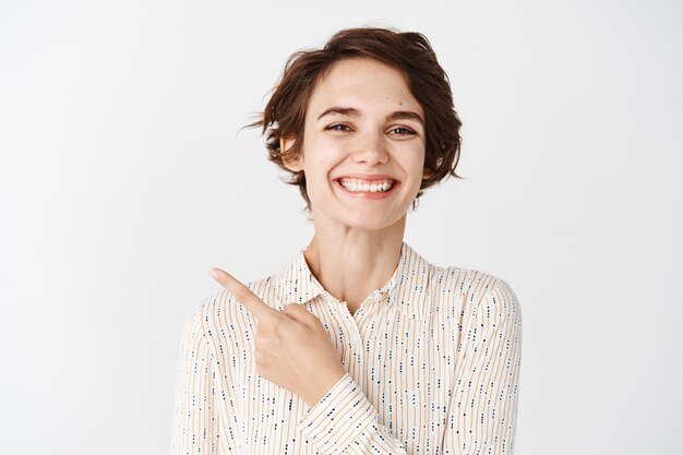 Portrait of caucasian woman in blouse smiling and pointing finger upper left corner, standing over white wall