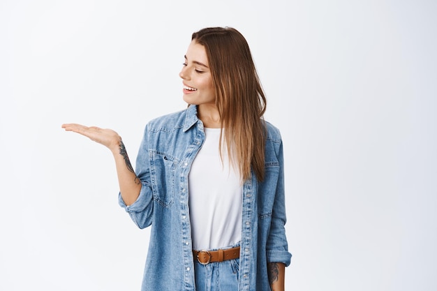 Portrait of caucasian young woman holding product on palm, looking at empty hand as if having something in it, smiling pleased, standing on white