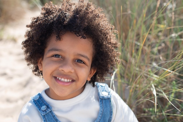 Free photo portrait of charming african american girl on beach. female model with curly hair in jeans clothes sitting on sand, looking at camera. portrait, beauty concept