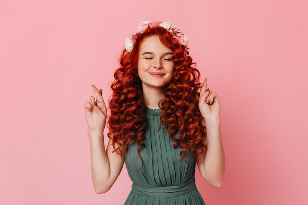 Portrait of charming lady in khaki dress smiling and crossing her fingers Girl with red curly hair smiles with closed eyes