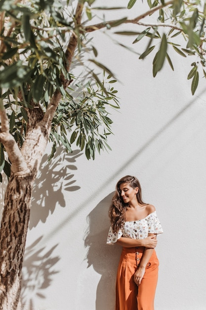 Free photo portrait of charming lady in summer resort outfit posing next to olive tree on white wall