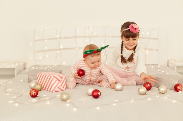 Portrait of charming sisters, cute little girls playing with Christmas balls on decorated bed with lights, children wearing casual attires, happy new year.