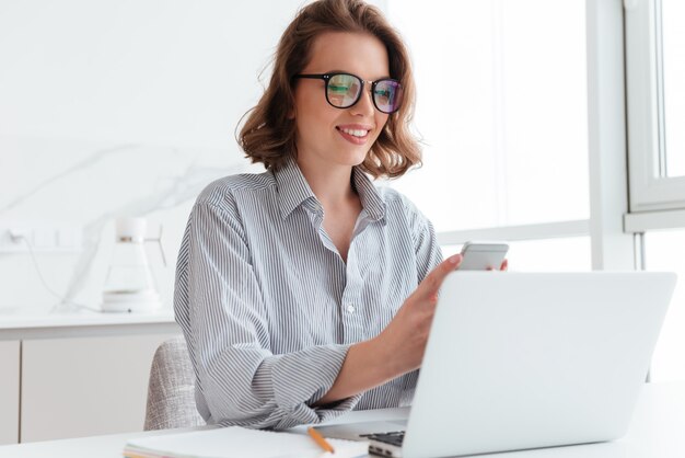 Portrait of charming woman in glasses and striped shirt using mobile phone while siting at workplace in white room