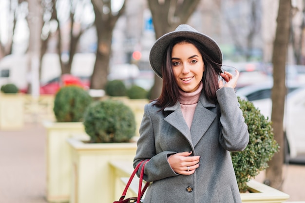 Portrait charming young fashionable woman in grey hat, coat walking on street in city park. Brunette hair, smiling, cheerful mood, elegant outlook.