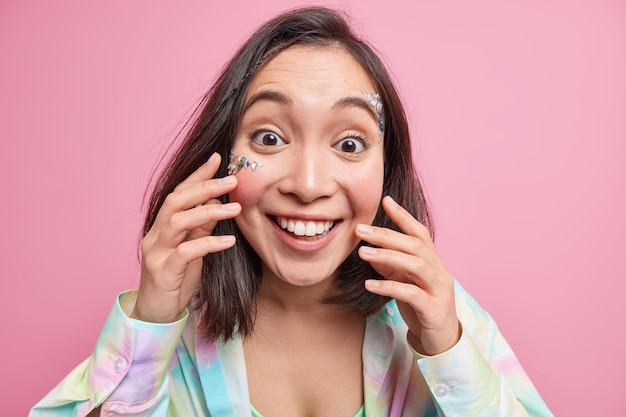 Portrait of cheeful Asian woman with toothy smile enjoys hearing positive news touches fresh skin has shiny stones pasted on face expresses happy authentic emotions isolated over pink wall