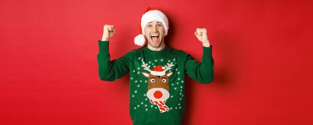 Portrait of cheerful attractive man celebrating new year wearing green sweater and santa hat shoutin...