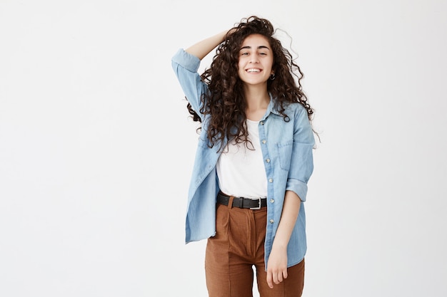 Portrait of cheerful brunette with long wavy hair, dressed in denim shirt and brown trousers, glad to recieve good news, Student girl smiles broadly, rejoices life, expresses positive emotions