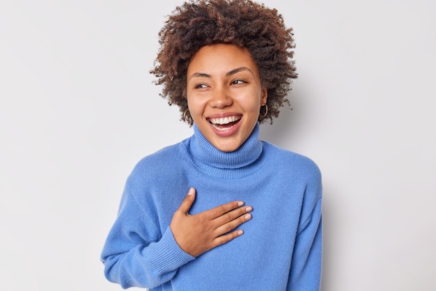 Portrait of cheerful curly haired woman smiles broadly keeps hand on chest cannot stop laughing at funny joke wears casual blue jumper isolated over white background. Positive human emotions concept