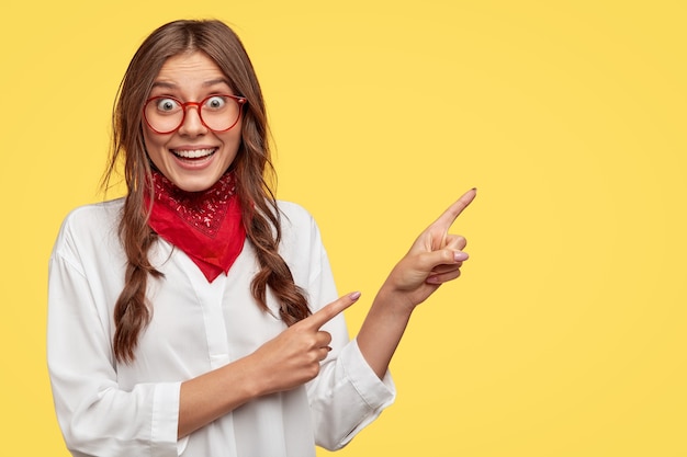 Portrait of cheerful European woman has two plaits, smiles broadly, wears stylish bandana and white shirt, points at upper right corner, models against yellow wall, suggests going at this place