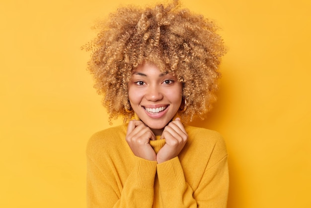 Free photo portrait of cheerful good looking woman smiles toothily keeps hands on collar of sweater looks directly at camera feels pleased poses against bright yellow background in studio. emotions concept