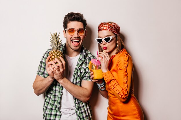 Portrait of cheerful guy in orange glasses holding pineapple and his girlfriend in satin dress drinking cocktail on white space.