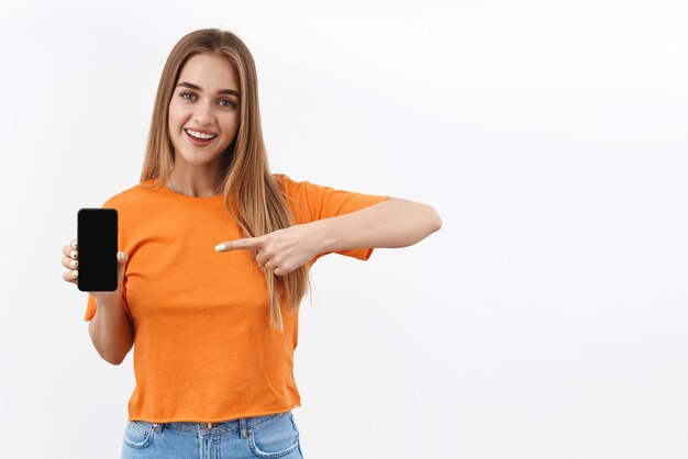 Portrait of cheerful, happy smiling blonde girl using mobile phone, pointing finger at smartphone display, showing shopping site