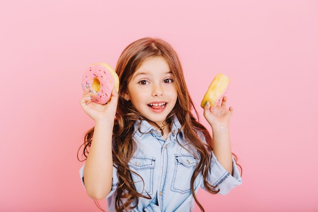 Portrait cheerful little girl with long brunette hair having fun to camera with colorful donuts isolated on pink background. Expressing true positive emotions of cute child. Place for text