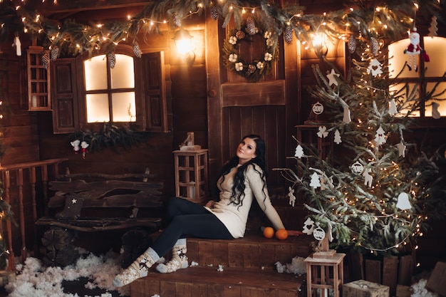 Portrait of cheerful and positive woman with long dark hair in sweater, jeans and warm socks holding wrapped gift for Christmas while sitting under decorated Christmas tree and snowfall