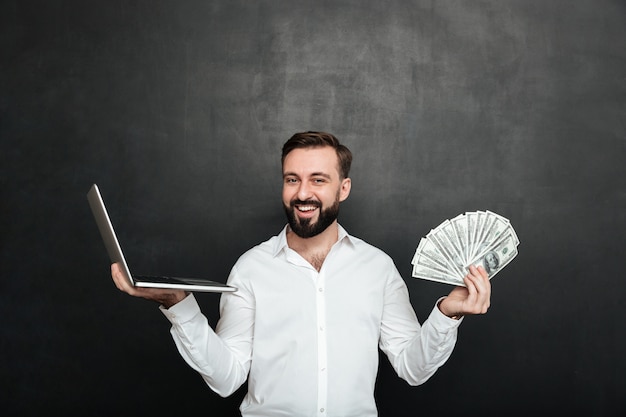 Portrait of cheerful rich man in white shirt winning lots of money dollar currency using his notebook over dark gray