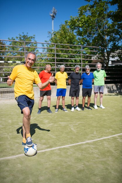 Portrait of cheerful senior man on football field. Team leader with gray hair in sport clothes standing, kicking ball, teammates in background. Football, sport, leisure concept