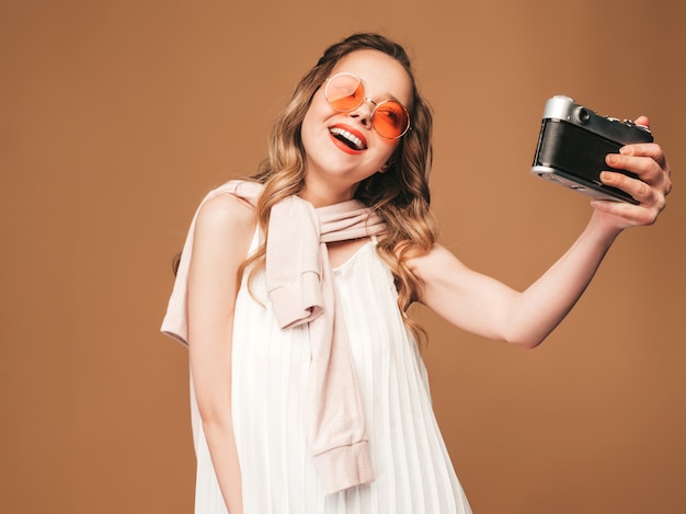 Portrait of cheerful smiling young woman taking photo selfie with inspiration and wearing white dress. Girl holding retro camera. Model in sunglasses posing