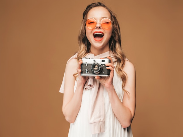 Free photo portrait of cheerful smiling young woman taking photo  with inspiration and wearing white dress. girl holding retro camera. model in sunglasses posing