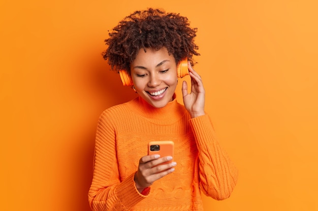 Free photo portrait of cheerful woman chooses song to listen from her playlist enjoys good sound in headphones concentrated in smartphone display dressed casually isolated over orange wall