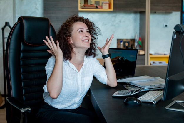 Portrait of cheerful young office worker woman sitting at office desk happy and excited with arms raised smiling cheerfully in office