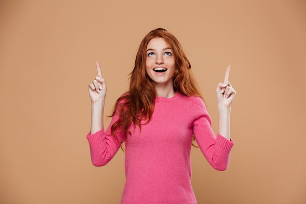 Portrait of a cheerful young redhead girl pointing up with fingers