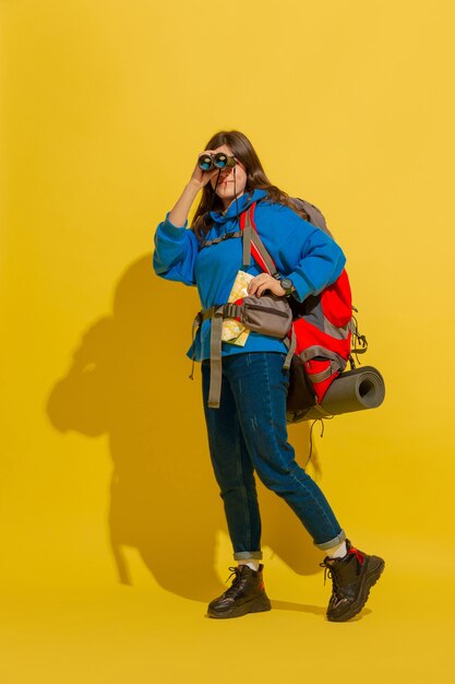 Portrait of a cheerful young tourist girl with bag and binoculars isolated on yellow studio wall