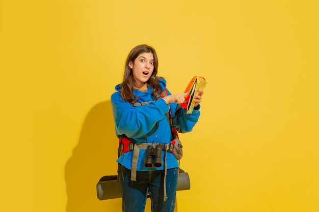 Portrait of a cheerful young tourist girl with bag and binoculars isolated on yellow studio wall