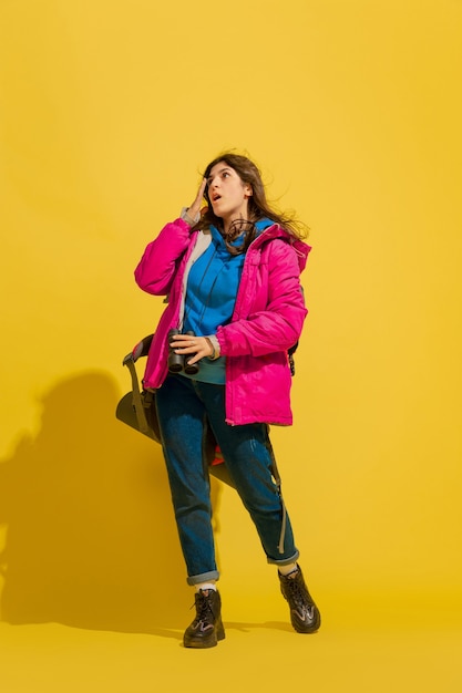 Portrait of a cheerful young tourist girl with bag and binoculars isolated on yellow studio wall