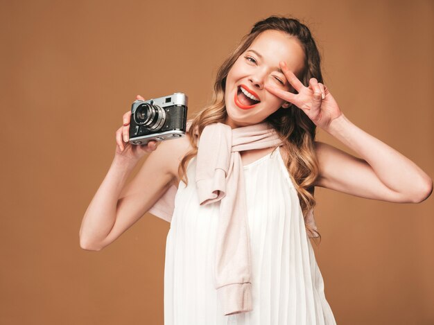 Portrait of cheerful young woman taking photo with inspiration and wearing white dress. Girl holding retro camera. Model posing