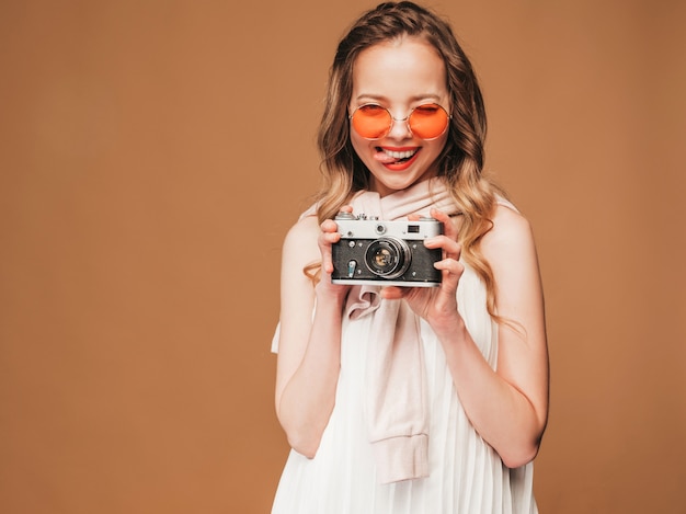 Free photo portrait of cheerful young woman taking photo with inspiration and wearing white dress. girl holding retro camera. model posing