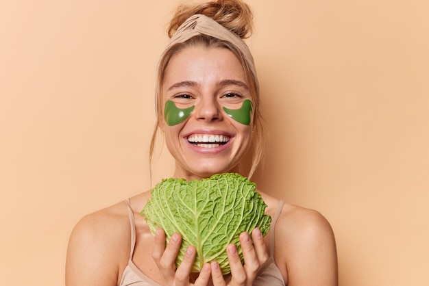 Portrait of cheerful young woman uses natural cosmetic and beauty products holds green fresh cabbage to put of skin for moisturising applies hydrogel patches under eyes stands bare shoulders