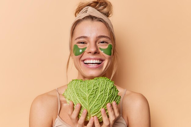 Portrait of cheerful young woman uses natural cosmetic and beauty products holds green fresh cabbage to put of skin for moisturising applies hydrogel patches under eyes stands bare shoulders