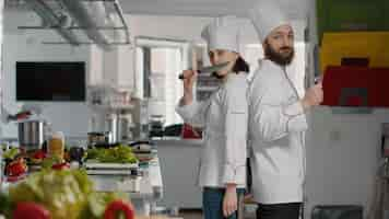 Free photo portrait of chefs team acting funny with knives in restaurant kitchen, making joke about professional cuisine service. cheerful man and woman working on gourmet meal dish with culinary recipe.